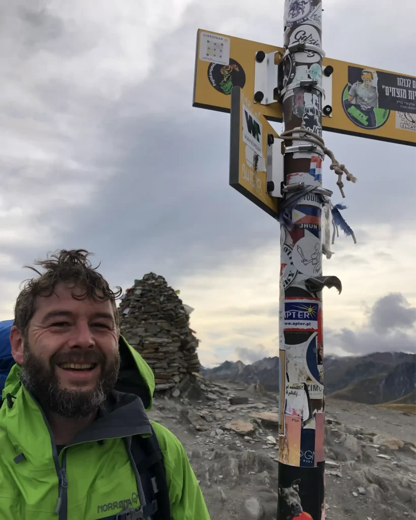 Tom at the Col de la Croix du Bonhomme on the Tour du Mont Blanc beside a sign covered with hikers' stickers, including a small Cheese Bar sticker.